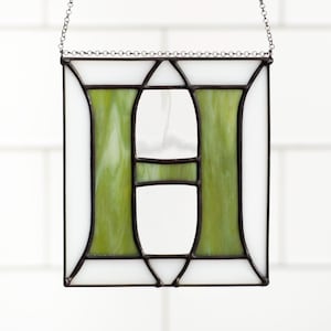 Stained glass panel featuring the letter "H" in a square frame. The letter is a vibrant green, bordered by white glass and black metal. A delicate chain suspends the artwork.