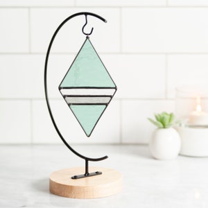 Geometric stained glass suncatcher made with green, grey and clear wavy textured glass. Made in the Tiffany Glass style with black patina. Displayed on an ornament hanger sitting on white marble in front of white subway tile and plant in background.