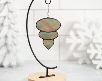 May include: A stained glass ornament in a teardrop shape, with three stacked sections of muted green, peach, and brown. The ornament hangs from a black metal stand with a wooden base, perfect for holiday decor.