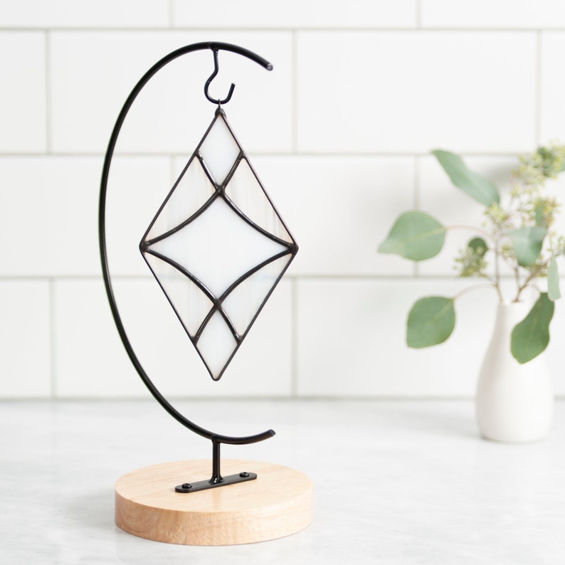 Curved design stained glass suncatcher made with opaque white and wispy white and clear glass with black patina. Displayed on an ornament hanger sitting on white marble in front of white subway tile and flowers in a vase in background.