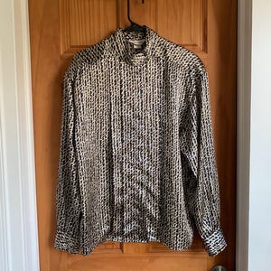 May include: A long-sleeved blouse with a high collar, featuring a repeating pattern of black and beige vertical lines. The blouse is hanging on a black hanger against a wooden door.