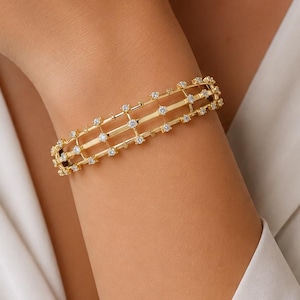 May include: A gold-toned cuff bracelet featuring a multi-strand grid design. The structure consists of three parallel horizontal bars connected by vertical segments, with small, clear, round stones set at each intersection point of the grid.