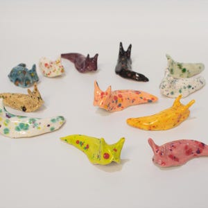 May include: A selection of ceramic slug ornaments in diverse colours and patterns. The slugs are in shades of yellow, pink, orange, blue, and white, with speckled and dotted designs. Some have pointed antennae.