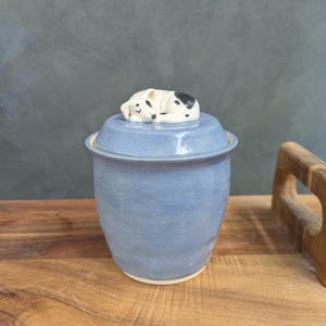 May include: A blue ceramic jar with a lid, featuring a small, sleeping dog figurine. The jar has a light blue glaze and a round shape. The dog figurine is white with black spots. The jar is on a wooden surface.