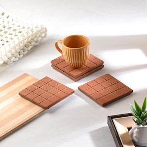 Puede incluir: Un juego de posavasos cuadrados de color terracota con un patrón de cuadrícula. Una taza a juego se encuentra encima de una pila de posavasos. La taza y los posavasos están sobre una superficie blanca.