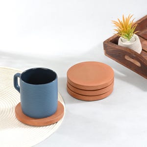 Op de afbeelding: Een donkerblauwe, geribbelde keramische mok staat op een ronde terracotta onderzetter. Daarnaast ligt een stapel van drie extra ronde terracotta onderzetters. De onderzetters hebben een gladde, matte afwerking en een roodbruine kleur.