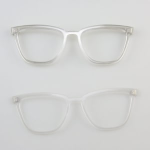 May include: Two pairs of clear, transparent eyeglasses are displayed against a white background. The top pair has a square frame, while the bottom pair has a slightly cat-eye shape. Both pairs are made of a clear plastic material.