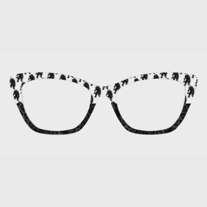 May include: Cat-eye style eyeglasses with a white frame featuring a repeating black Bigfoot pattern. The lower portion of the frame is black with a speckled design. The lenses are clear.
