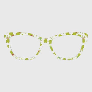 May include: Cat-eye glasses featuring a white frame with a repeating pattern of small green frogs. Each frog is depicted in a simple line drawing style with a small pink or blue circular detail on its body, distributed across the entire frame.