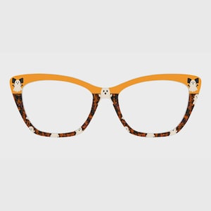 May include: Cat-eye glasses featuring a solid orange upper frame and a lower frame patterned with small white ghosts and black bows against a dark, mottled background. A small white ghost is centered on the bridge of the frame.