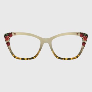 May include: Cat-eye spectacles with a cream-coloured frame and a tortoiseshell pattern on the lower section. The outer corners feature red rose floral designs. The spectacles have clear lenses.