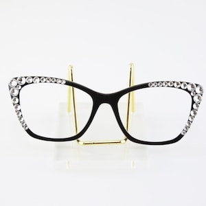 May include: Black cat-eye eyeglasses with clear lenses, embellished with sparkling rhinestones along the top frame. The glasses are displayed on a gold-colored stand against a white background.