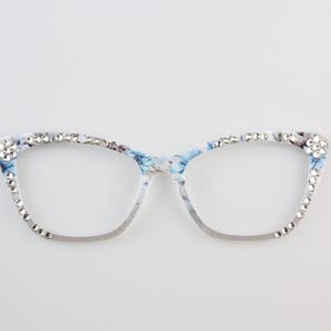 Op de afbeelding: Bril in cat-eye stijl met een bloemenpatroon in blauw en wit. De monturen zijn versierd met sprankelende strass-steentjes langs de boven- en buitenranden. Een modieus accessoire.
