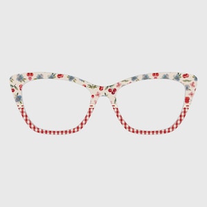 May include: Cat-eye eyeglasses with a cream-colored floral pattern featuring red cherries and blue flowers. The lower portion of the frames has a red and white gingham pattern, creating a vintage-inspired look.