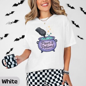 May include: A white t-shirt featuring a graphic of a black animal paw stirring a purple cauldron filled with light blue bubbling liquid. The text Paws & Potions is printed on the cauldron. A black mug with a white ghost pattern is also visible.