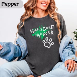 May include: A charcoal grey crewneck sweatshirt featuring a green paint splatter graphic with the white text 'HARDCORE BARKOUR' and a white paw print icon. The garment is layered under a light blue denim jacket, creating a casual layered look.