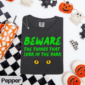 May include: A charcoal gray t-shirt featuring the bright green text BEWARE THE THINGS THAT LURK IN THE DARK above a pair of yellow cat eyes. The shirt has a crew neckline and a Comfort Colors brand label visible on the inside of the collar.