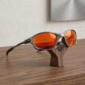Op de afbeelding: Grijze zonnebril met oranje glazen op een houten displaystandaard. De zonnebril heeft een gebogen montuur en donkergrijze pootjes. De standaard is een donkerbruine, gestileerde Y-vorm, die op een houten oppervlak staat.