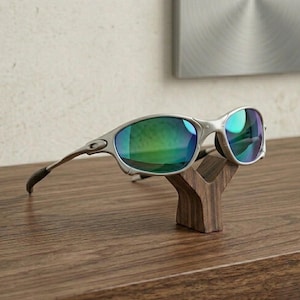 May include: Silver-framed sunglasses with green-tinted lenses rest on a small, dark wooden display stand. The sunglasses have a sleek, modern design, and the stand is shaped like a stylized letter "Y". The background is a neutral color.