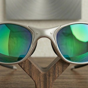 May include: Silver-framed sunglasses with green and blue reflective lenses. The sunglasses are displayed on a wooden stand. The lenses have a gradient effect, transitioning from green to blue. The frame has a matte finish.