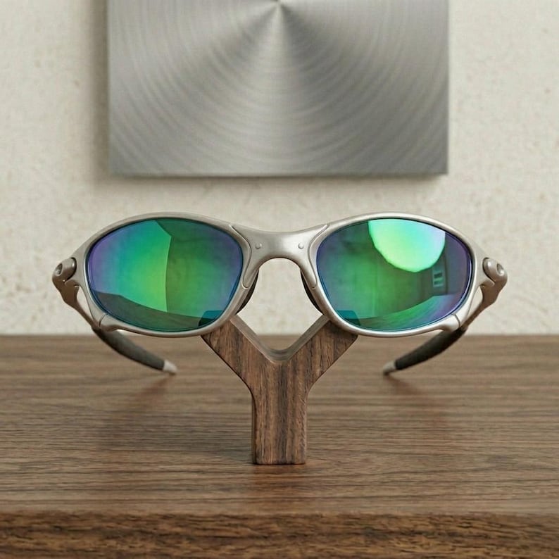 May include: Silver-framed sunglasses with green-tinted lenses rest on a wooden display stand. The sunglasses have a sleek, modern design. The stand is a dark brown, Y-shaped structure, placed on a wooden surface.