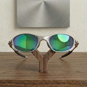 May include: Silver-framed sunglasses with green-tinted lenses rest on a wooden display stand. The sunglasses have a sleek, modern design. The stand is a natural wood color, and the background features a metallic wall hanging.