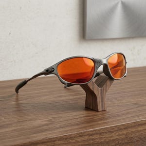 Op de afbeelding: Grijze zonnebril met oranje glazen op een houten displaystandaard. De zonnebril heeft een gebogen montuur en donkergrijze pootjes. De standaard is een donkerbruine, gestileerde Y-vorm, die op een houten oppervlak staat.
