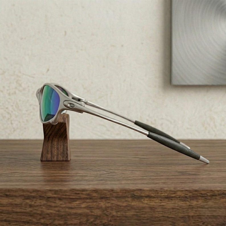 May include: Silver-framed sunglasses with iridescent lenses are displayed on a wooden stand. The lenses reflect shades of green and blue. The temples are thin and extend to black tips. The sunglasses are set against a neutral background.