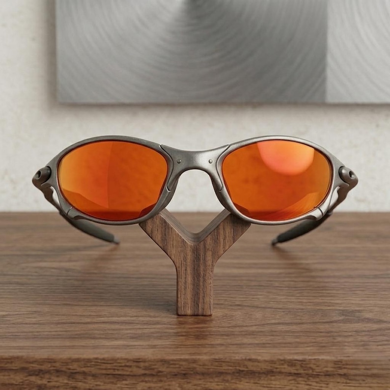 Op de afbeelding: Een zonnebril met een grijs montuur en oranje glazen. De bril wordt getoond op een houten standaard. De glazen reflecteren licht en het montuur heeft een matte afwerking. De zonnebril is bedoeld voor oogbescherming.