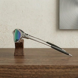 May include: Silver-framed sunglasses with mirrored lenses displaying shades of green and blue. The sunglasses are resting on a small, dark wooden stand. The temples are thin and extend to black tips.
