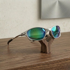 May include: Silver-framed sunglasses with green-tinted lenses rest on a small, dark wooden display stand. The sunglasses have a sleek, modern design, and the stand is shaped like a stylized letter "Y". The background is a neutral color.