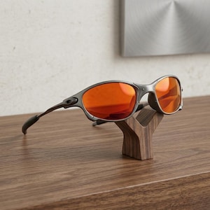 Op de afbeelding: Grijze zonnebril met oranje glazen op een houten displaystandaard. De zonnebril heeft een gebogen montuur en donkergrijze pootjes. De standaard is een donkerbruine, gestileerde Y-vorm, die op een houten oppervlak staat.