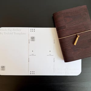 Może przedstawiać: Biały szablon do składania B5 marki Black Fig Atelier z nadrukowanymi prowadnicami i kwadratami testowymi o boku 1 cala, obok ciemnobrązowego notatnika o fakturze drewna, zabezpieczonego cienką gumką i małą zawieszką w kolorze złotym.