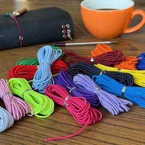Könnte beinhalten: Eine Sammlung bunter Gummischnüre in verschiedenen Farbtönen, darunter Rosa, Grün, Blau und Lila, sind ordentlich gebündelt. Ein schwarzes Notizbuch, eine orangefarbene Tasse mit Kaffee und ein Stift sind ebenfalls auf einer Holzoberfläche zu sehen. Die Schnüre sind wahrscheinlich zum Basteln.