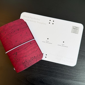 May include: A deep red cork bifold wallet with black flecks and a white elastic band, positioned next to a white rectangular template labeled Block Fig Atelier Pocket Bifold Template with a 1 inch test square and eyelet placement markings.