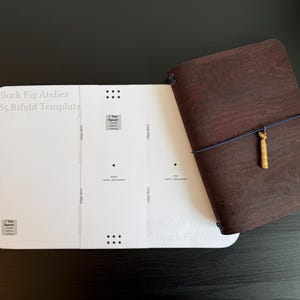 Op de afbeelding: Een donkerbruin kurken notitieboek met een blauwe elastische band en een goudkleurige bedel, geplaatst naast een wit papieren sjabloon met het label Block Fig Atelier B5 Bifold Template, een testvierkant van 2,54 cm en uitlijning.