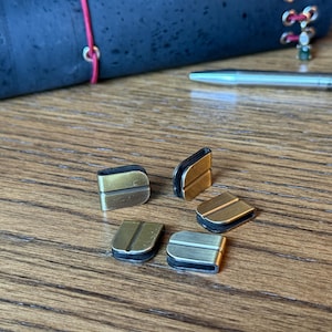 May include: Five small, rectangular metal clasps with a gold and silver finish are scattered on a wooden surface. A dark blue, textured object and a silver pen are in the background. The clasps have a curved design.