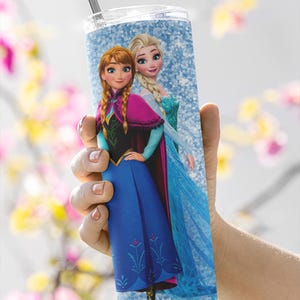 May include: A tall, cylindrical tumbler featuring a design of two animated female characters, Anna and Elsa, from the movie Frozen. The tumbler has a silver straw and a clear lid. The background is a light blue glitter pattern. The characters are in their signature outfits.