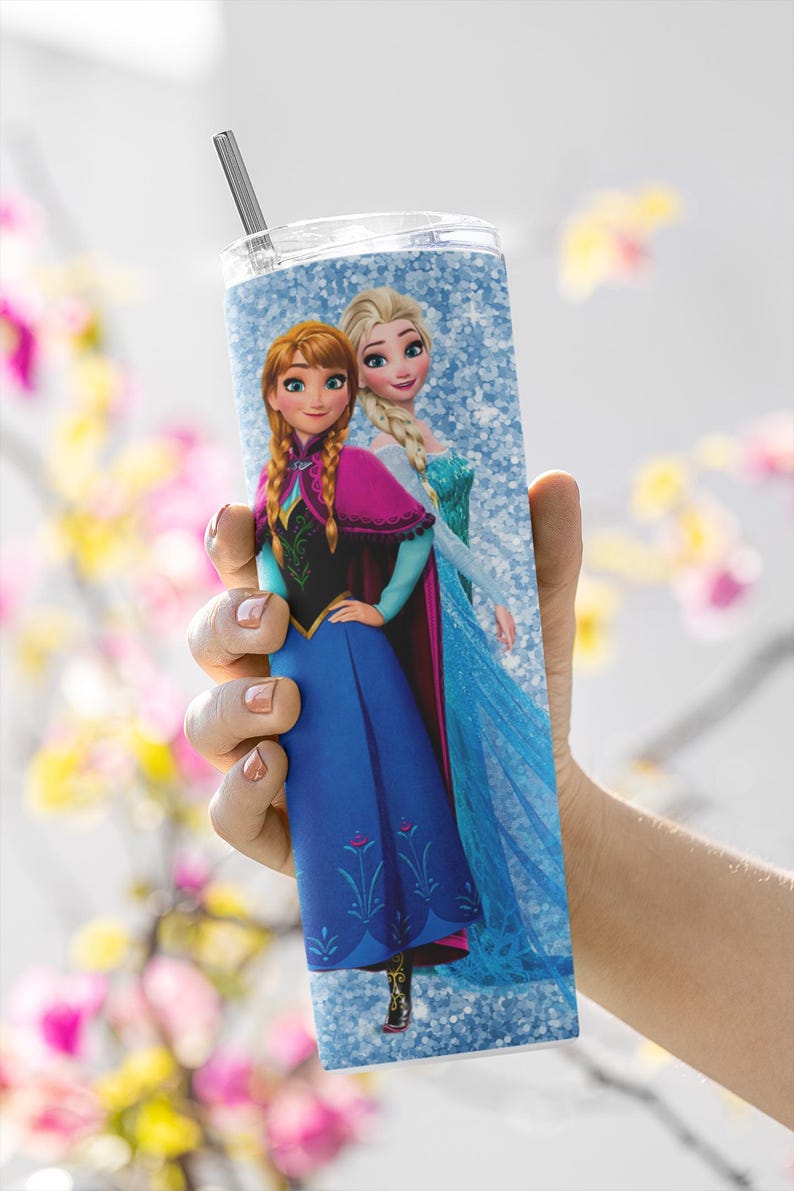May include: A tall, cylindrical tumbler featuring a design of two animated female characters, Anna and Elsa, from the movie Frozen. The tumbler has a silver straw and a clear lid. The background is a light blue glitter pattern. The characters are in their signature outfits.