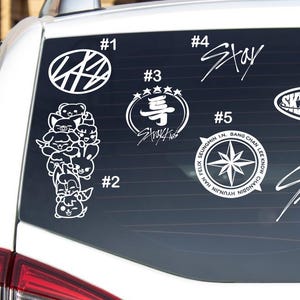 May include: White vinyl decals on a car window. The decals include the text "Stay", "Stray Kids", and other graphics. The decals are numbered and include a compass rose, a star, and cartoon animal characters.
