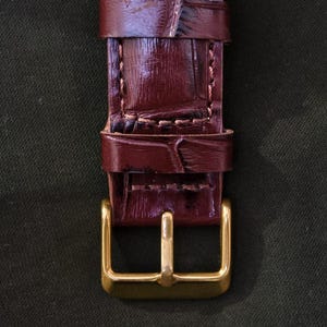 May include: Close-up of a burgundy leather watch strap with a gold-colored buckle. The strap has a textured, crocodile-like pattern and is stitched with a lighter thread. The buckle is rectangular with a single bar.
