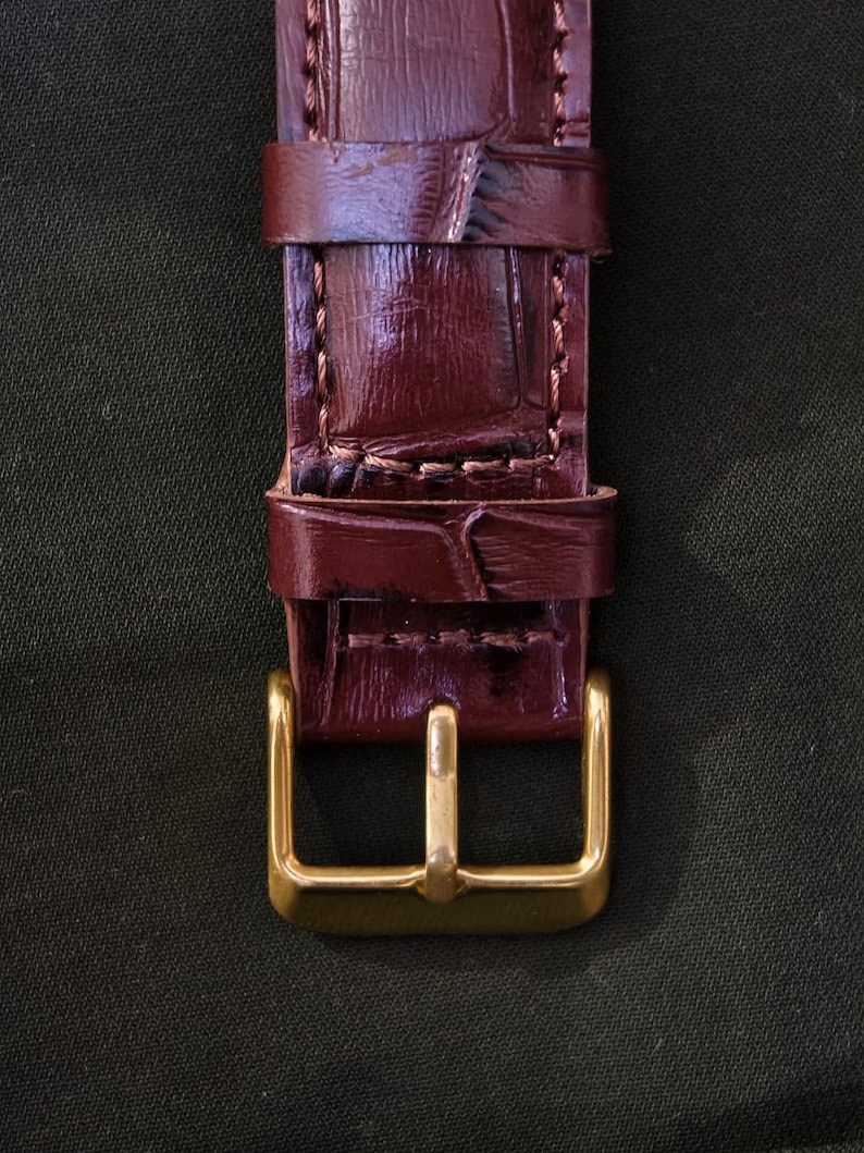 May include: Close-up of a burgundy leather watch strap with a gold-colored buckle. The strap has a textured, crocodile-like pattern and is stitched with a lighter thread. The buckle is rectangular with a single bar.