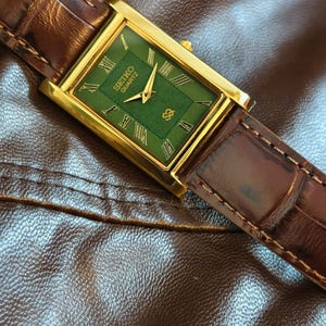 Może przedstawiać: Złoty zegarek Seiko Quartz z prostokątną zieloną tarczą i brązowym sk&oacute;rzanym paskiem. Tarcza zegarka posiada rzymskie cyfry i logo Seiko. Zegarek jest umieszczony na brązowej sk&oacute;rzanej powierzchni.