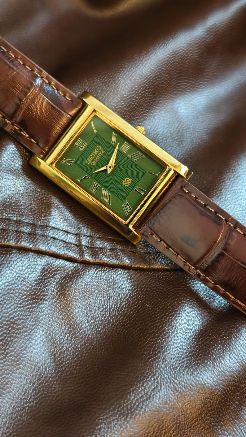 Może przedstawiać: Złoty zegarek Seiko Quartz z prostokątną zieloną tarczą i brązowym sk&oacute;rzanym paskiem. Tarcza zegarka posiada rzymskie cyfry i logo Seiko. Zegarek jest umieszczony na brązowej sk&oacute;rzanej powierzchni.