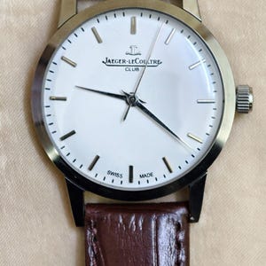 May include: A silver-toned Jaeger-LeCoultre Club watch with a white face and black hands. The watch has a brown leather band. The face displays the Jaeger-LeCoultre logo and the words "SWISS MADE".