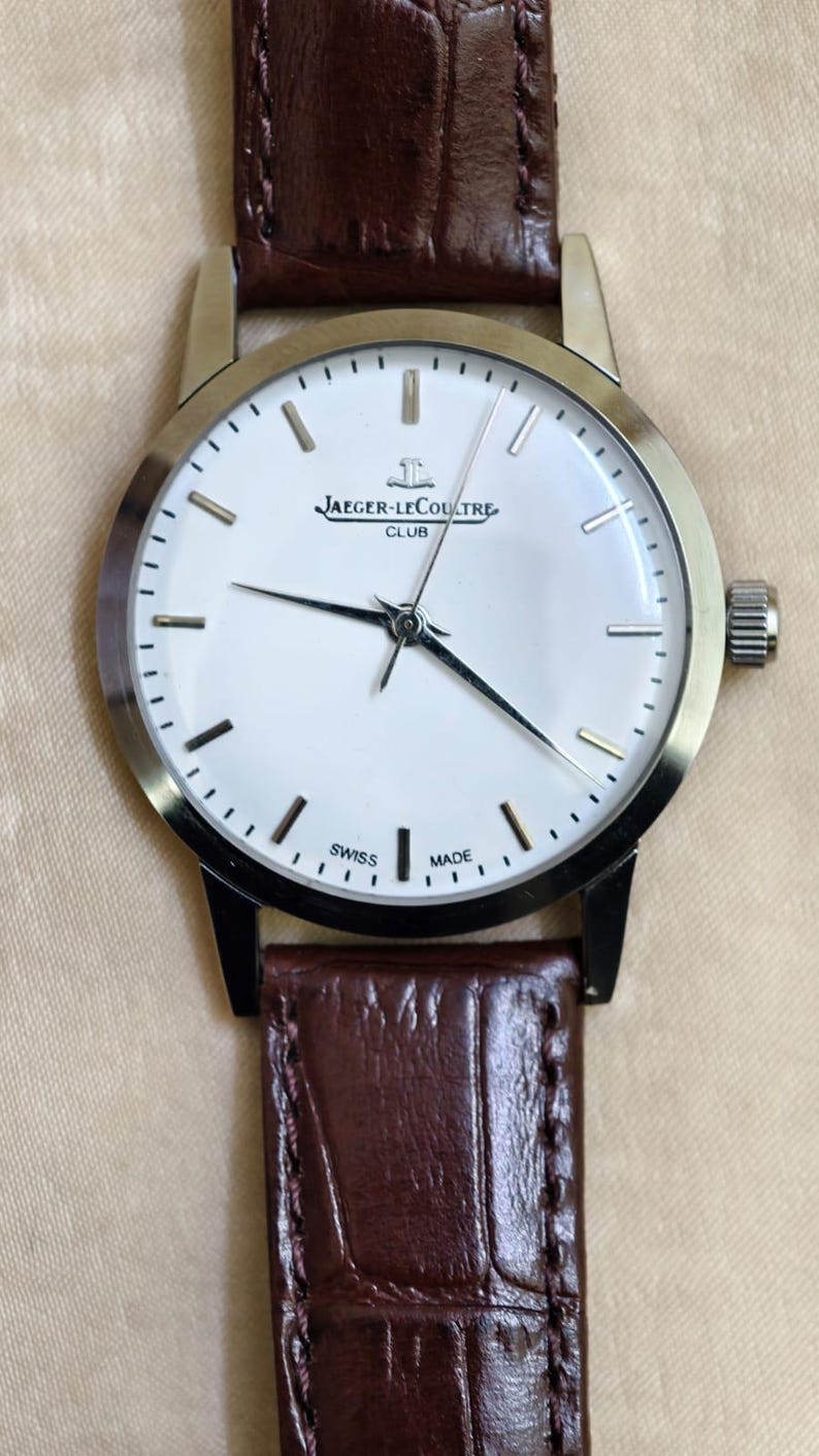 May include: A silver-toned Jaeger-LeCoultre Club watch with a white face and black hands. The watch has a brown leather band. The face displays the Jaeger-LeCoultre logo and the words "SWISS MADE".