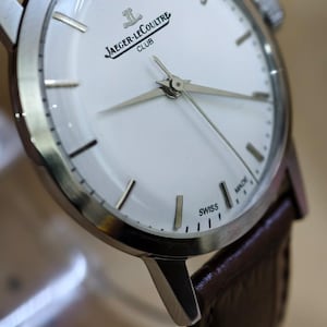 May include: A silver-toned Jaeger-LeCoultre Club watch with a white face and brown leather strap. The watch features silver hands and markers, with the brand name and "SWISS MADE" text visible on the face.