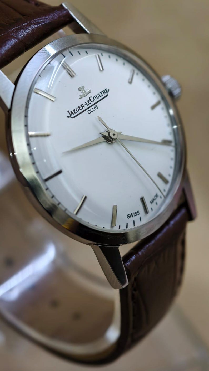 May include: A silver-toned Jaeger-LeCoultre Club watch with a white face and brown leather strap. The watch features silver hands and markers, with the brand name and "SWISS MADE" text visible on the face.