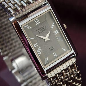Puede incluir: Un reloj Seiko Quartz rectangular con una correa de metal plateado tejido. La esfera del reloj es gris con números romanos y agujas blancas. El reloj tiene una caja plateada y una esfera gris texturizada.