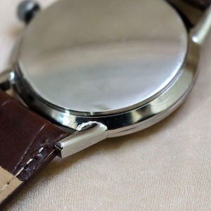 May include: Close-up of a silver wristwatch with a brown leather strap. The watch face is not visible, but the polished metal back and the strap's texture are prominent. The watch's lugs are visible.
